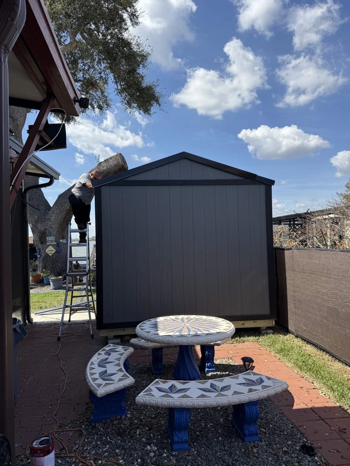 Gable-end exterior view of a 10 by 16-foot Patriot shed beside a screened patio, with dark trim and a crew member working along the roof edge
