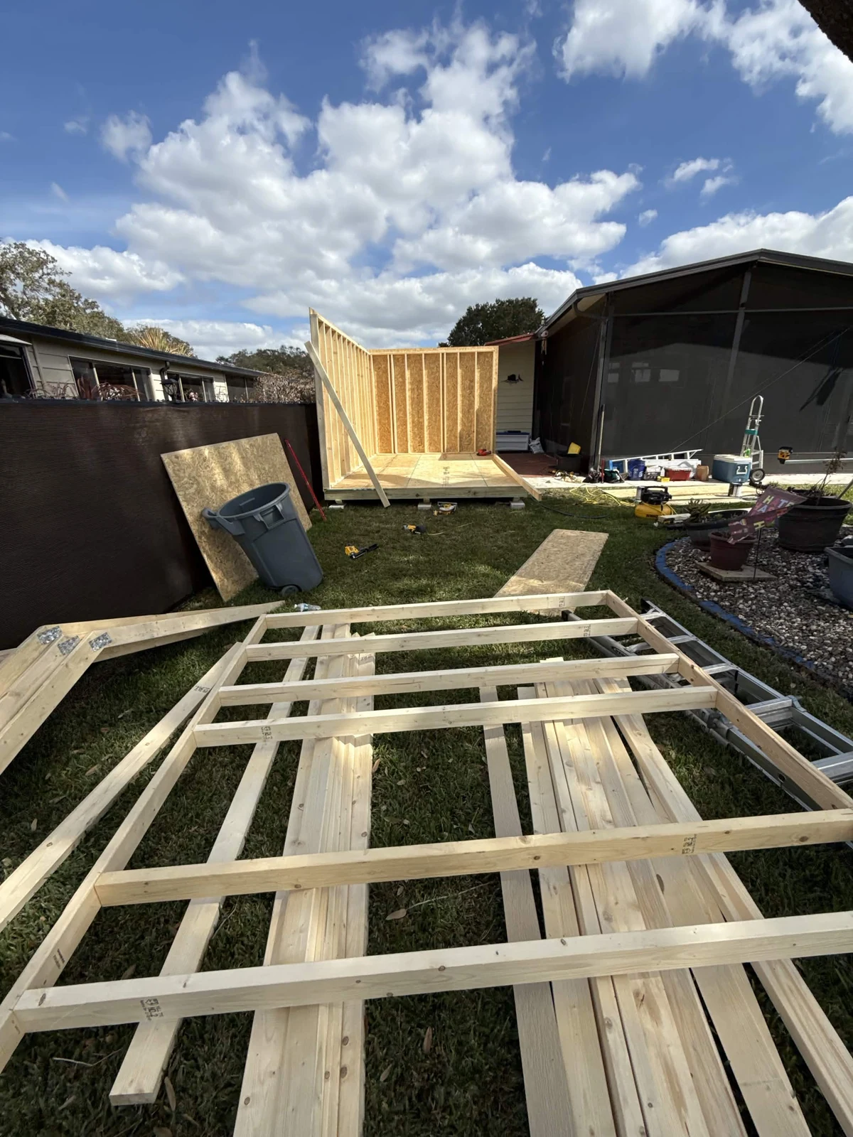 Early construction of the 10 by 16-foot shed, showing a floor platform, standing walls, and a framed wall section laid out in the yard